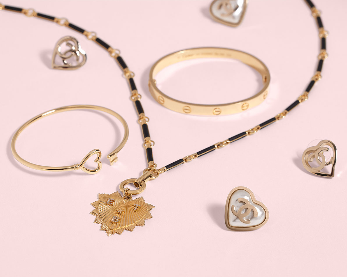 One Yellow Gold Cartier Love Bracelet laying next to one Tiffany Yellow Gold Wire Heart Key Bracelet, two pairs of Chanel heart earrings, and one Foundrae Yellow Gold Diamond Black Onyx Oversized Heart Love Token Medallion Hanging Clockweight Necklace 