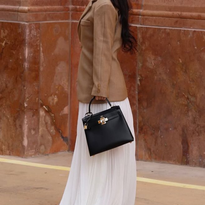 a woman in a white long skirt and brown blazer holding a black Hermes Kelly bag