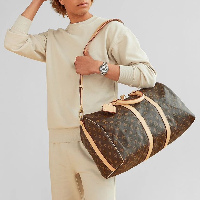 a man wearing a beige sweatsuit and Rolex Panda watch holding a Louis Vuitton Monogram Keepall bag