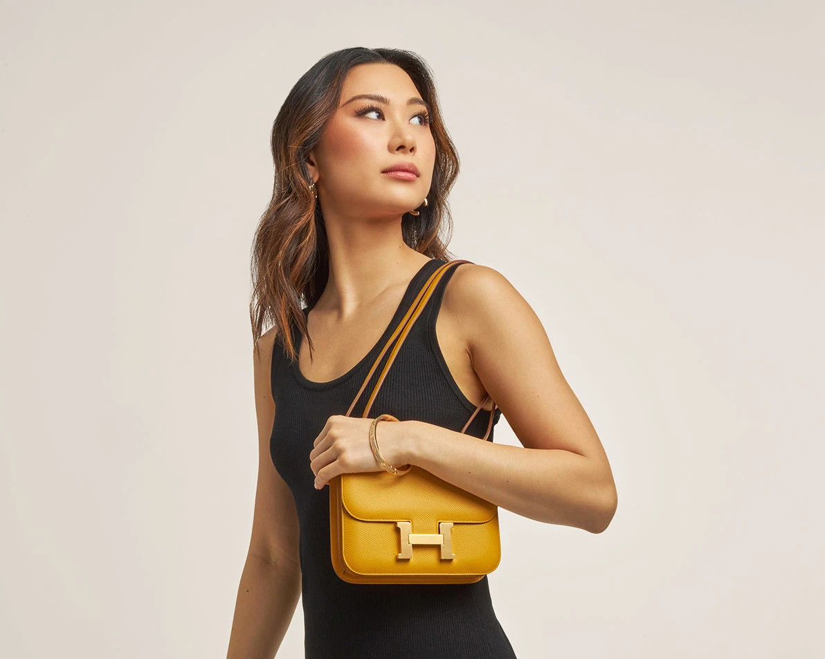 a woman wearing a black sleeve dress and holding a yellow Hermes Constance 18 size bag