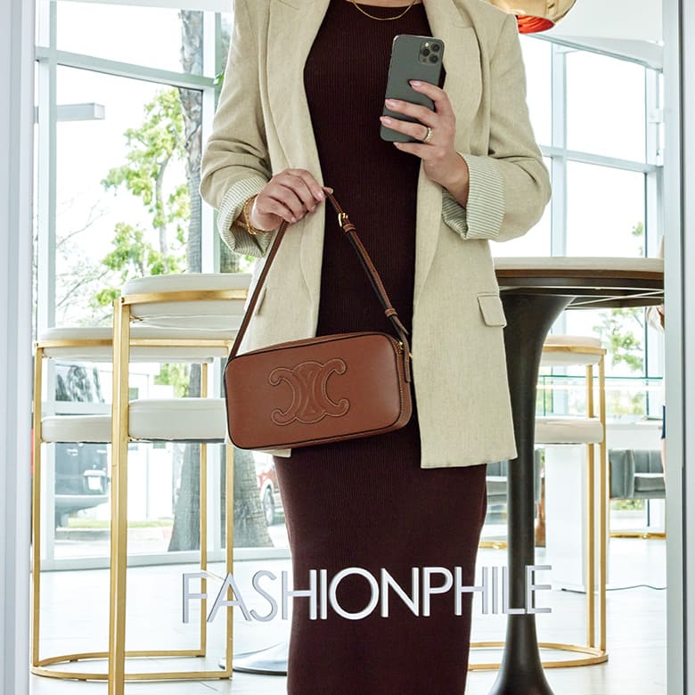 Woman wearing a black dress and beige blazer holding a brown Celine Camera bag taking a selfie in a mirror at the Carlsbad FASHIONPHILE studio. The mirror reads "FASHIONPHILE" in white.