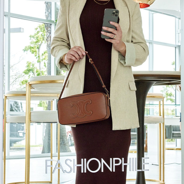 Woman wearing a black dress and beige blazer holding a brown Celine Camera bag taking a selfie in a mirror at the Carlsbad FASHIONPHILE studio. The mirror reads "FASHIONPHILE" in white.