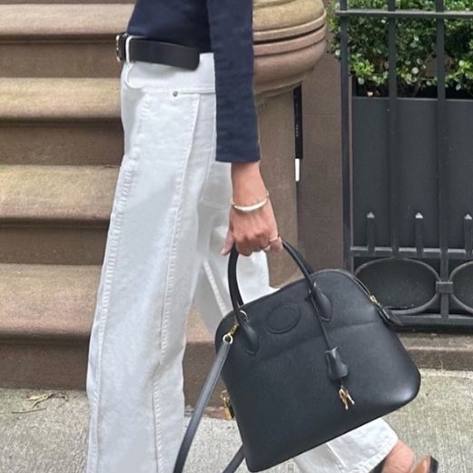 A woman in a navy sweater and white pants holding a black Hermes Bolide bag