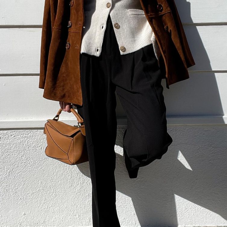 A woman in white cardigan, brown suede jacket and black slacks holding a brown Loewe Puzzle bag