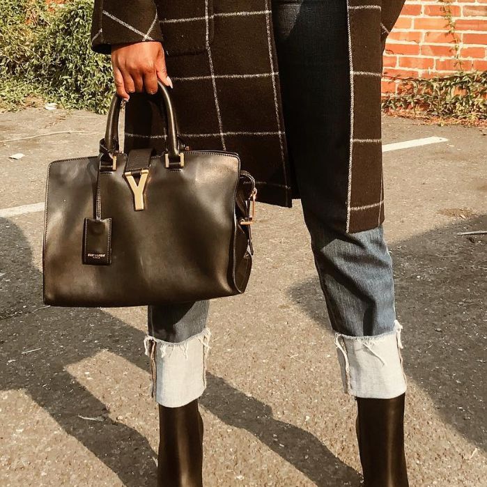 a woman in a plaid black coat and denim jeans holding a black Saint Laurent Cabas bag