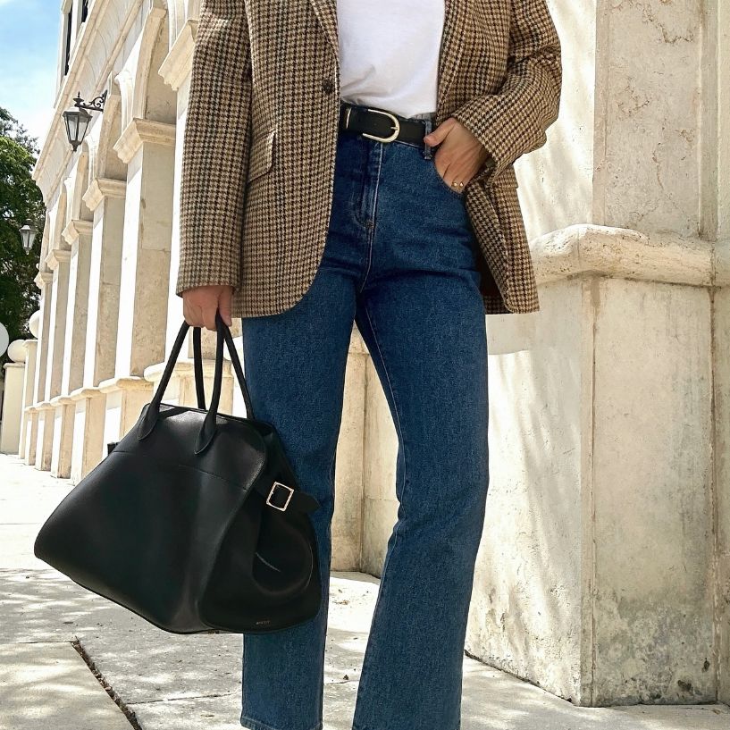 A woman in denim pants and a beige blazer holding a black The Row Margaux