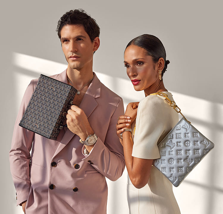 a woman in an ivory color dress holding a silver Louis Vuitton Coussin bag with a gold shoulder chain and a man in a mauve color suit holding a small Goyard trunk bag