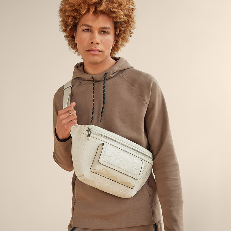 a man wearing a brown hooded sweatshirt and holding a white monogram Gucci belt bag