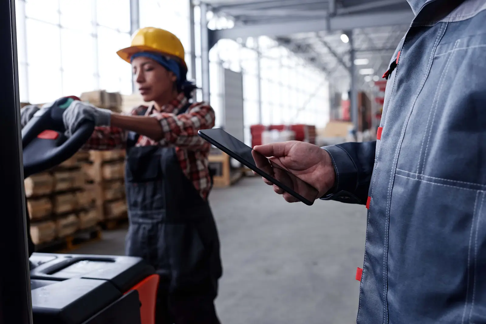 Warehouse worker using a rugged tablet while forklift operates in background