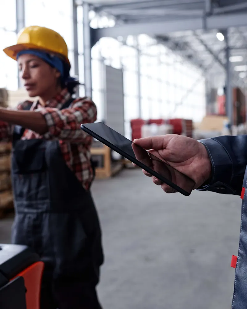 Warehouse worker using a rugged tablet while forklift operates in background
