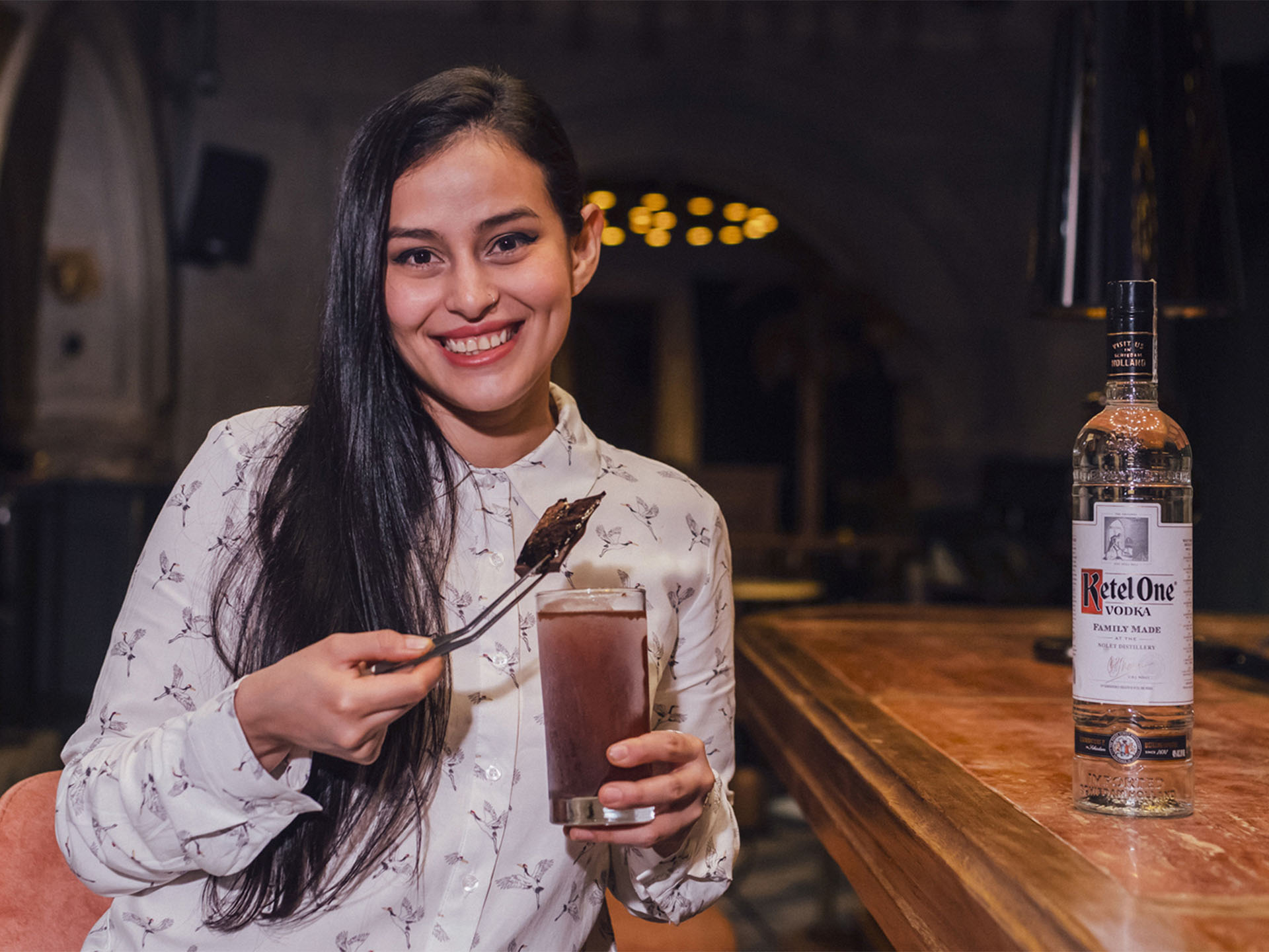 Dianna Alejandra Bernal tenant un cocktail dans un bar avec une bouteille de vodka Ketel One