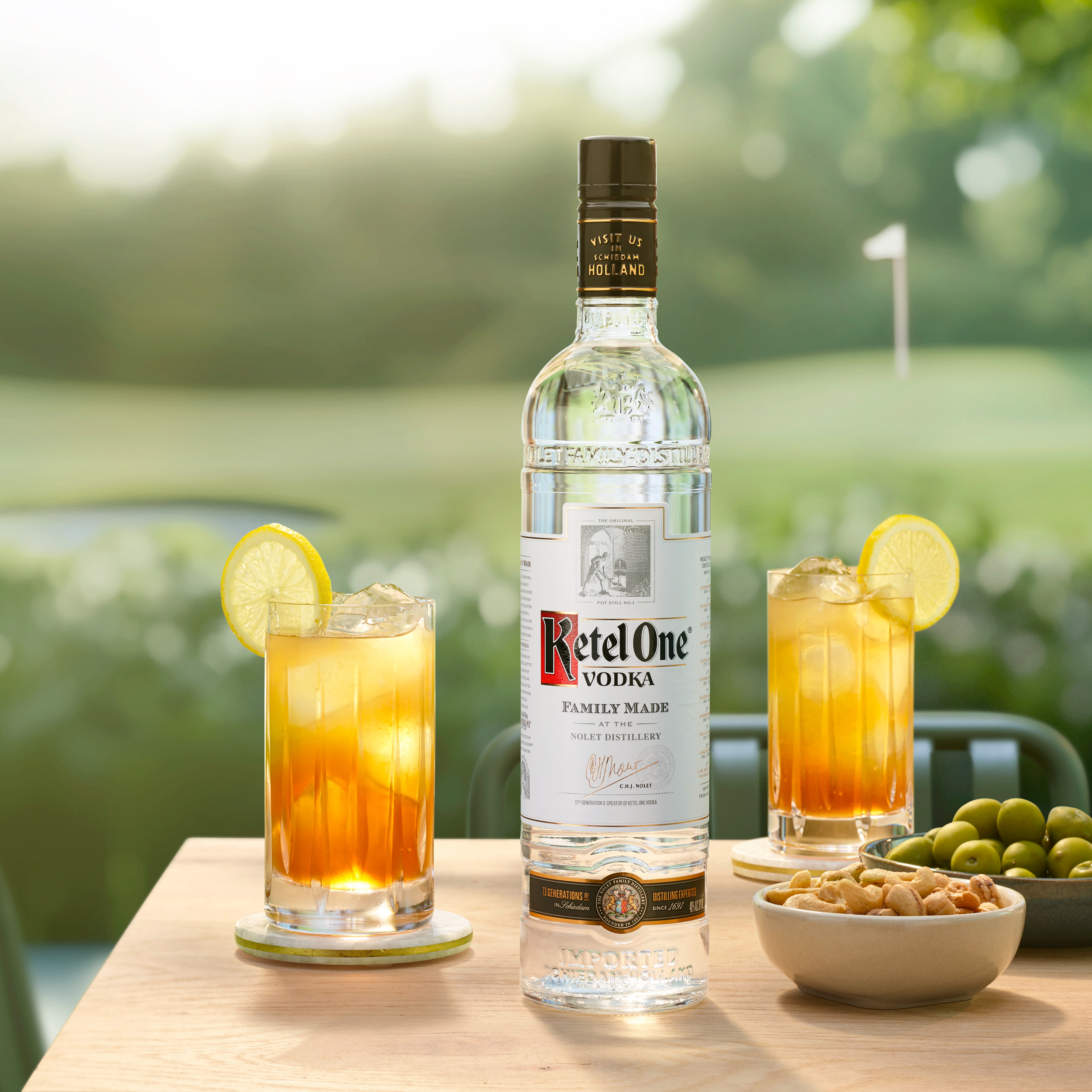 Bottle of Ketel One vodka on a table with two glasses of Tee Time cocktail, a bowl of nuts and a bowl of olives. In the background is a golf course.