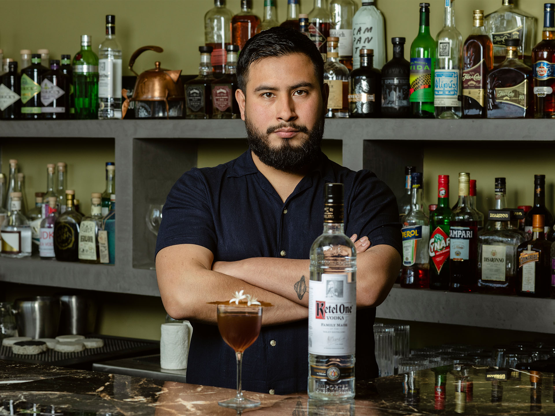 Luis Alza with his arms folded standing in a bar with Ketel One Vodka and a cocktail