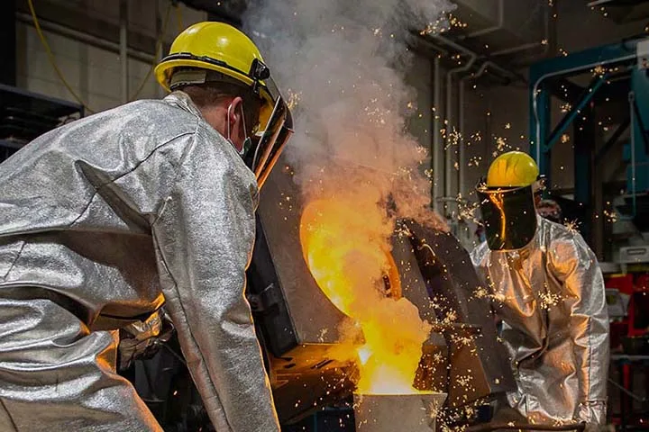 Two people in protective gear and hard hats pouring hot liquid into container