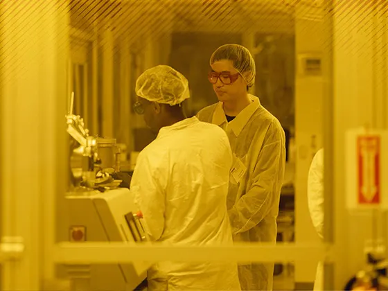 Two students working in a clean room wearing hair nets and lab coats.