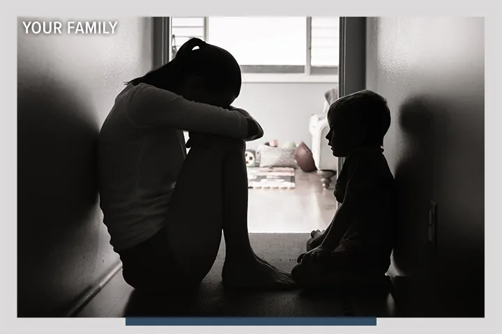 A mother crouched over in distress in a dark hallway with her child. There is an overlay that says Your Family.