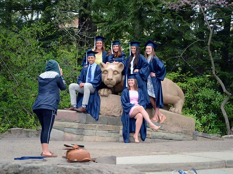 A wide photo of the Nittany Lion Shrine area. There is a woman in the foreground taking a photo of six students sitting around the Lion in their navy caps and gowns for graduation.