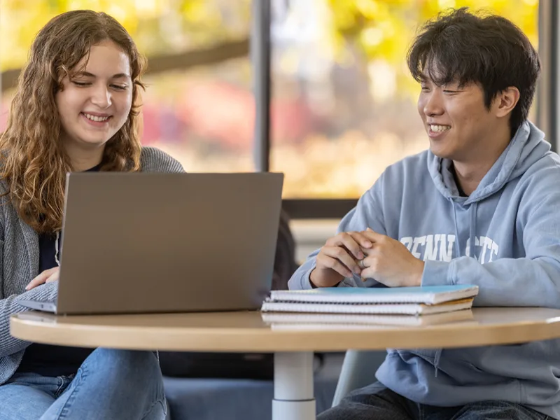 Two Penn State students sitting at a table on campus and working on a laptop.