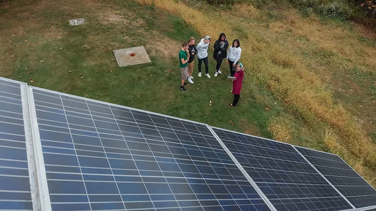 Photo of students and an instructor in a grass field looking up at a solar panel