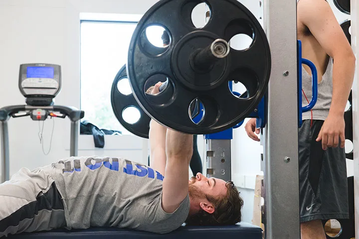 Photo of a student bench pressing in campus gym while friend assists