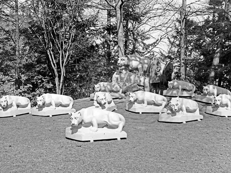 A black and white photo of two men (barely seen) standing next to the Lion Shrine. In front are 10 smaller replicas of the shrine.