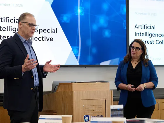 Kevin Linderman, dressed in a black suit, and Donna Palumbo, in a blue blazer and black pants stand at the front of a conference room with graphics projected behind them.