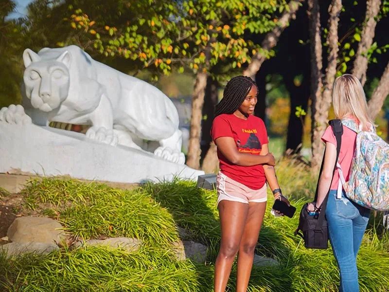 A white, stone lion shrine looks directly at the camera. It sits atop stones in front of a green lawn and students in the foreground.