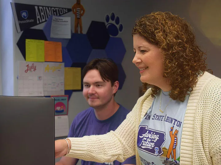 Two people in front of a computer, both are smiling while one gestures towards the computer. 