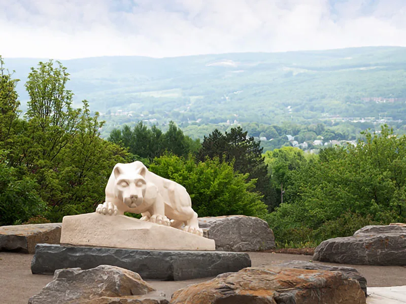 In the right corner, the Nittany Lion shrine faces away from the camera and towards the left. Students pat the front of the statue, and there is a Penn State Lehigh Valley building visible in the background.