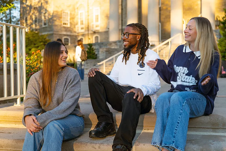 3 students in discussion enjoying a sunny day on the exterior steps of a campus building. 