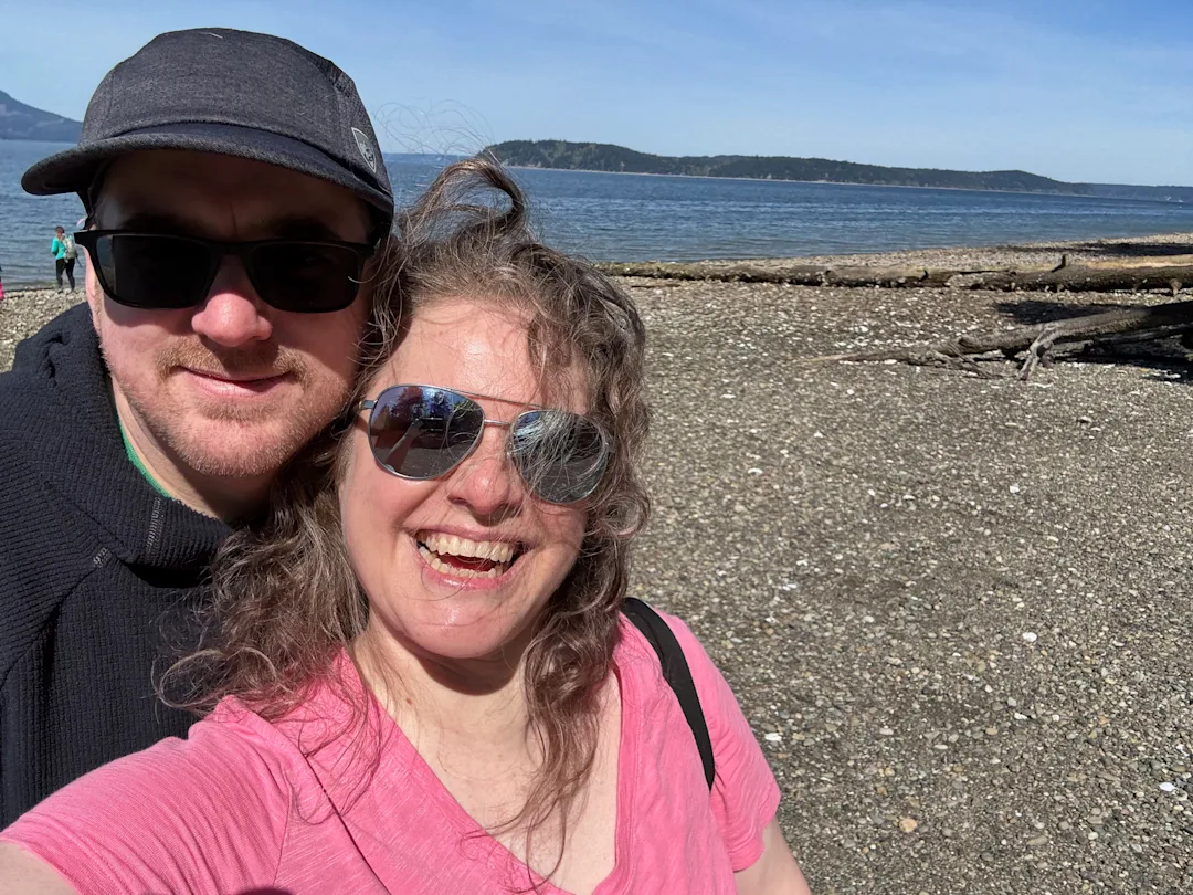 Two people with sunglasses standing on a beach beside a large body of water