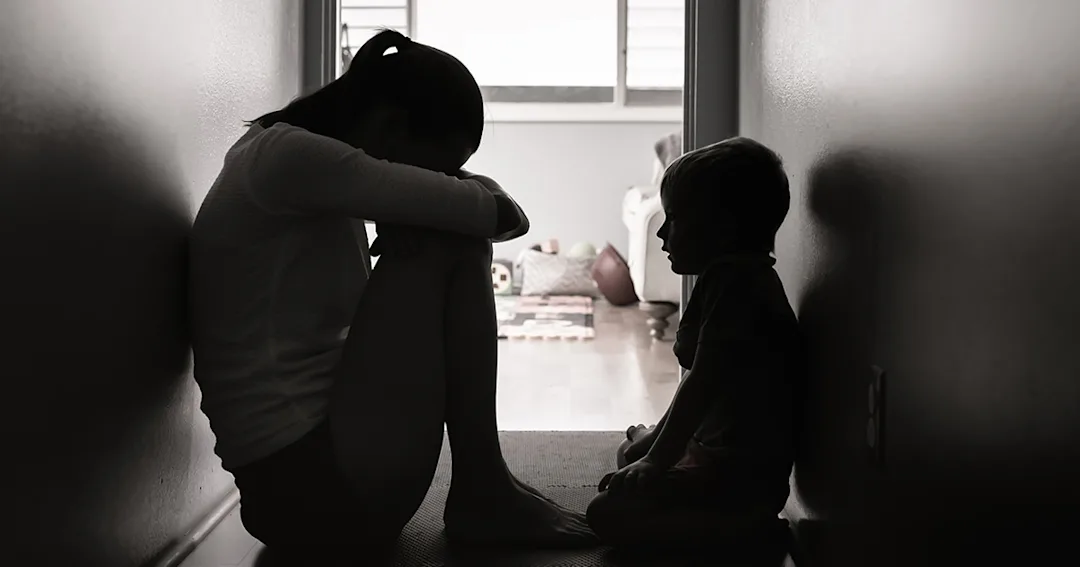 A mother crouched over in distress in a dark hallway with her child