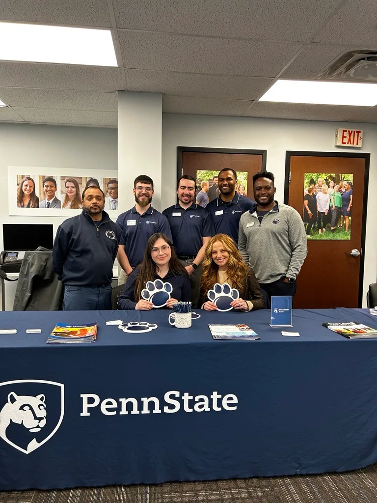 Seven Penn State staff member smiling behind table with Penn State navy table cover