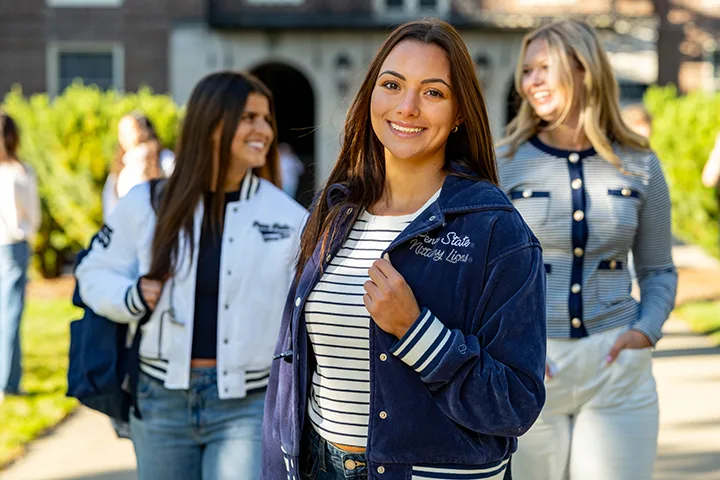 Female student proudly displaying her Penn State Jacket as she walks across a sunlit campus.