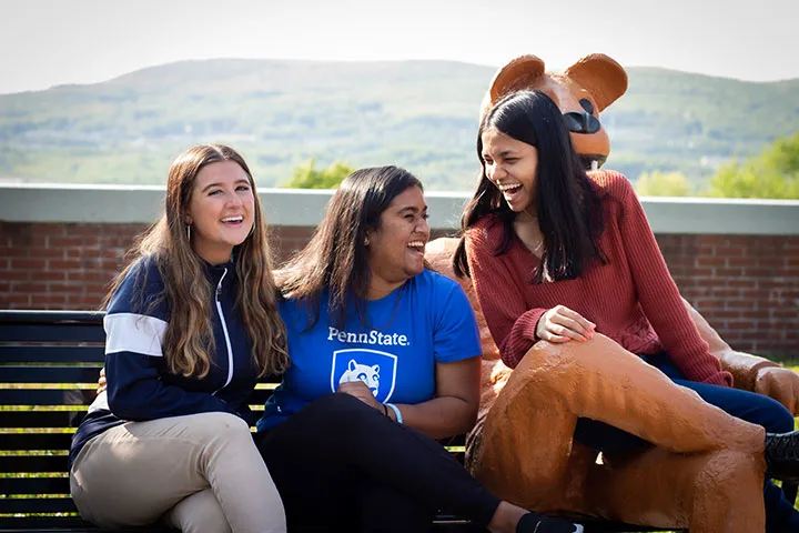 Photo of students laughing and sitting on a bench with the Nittany Lion mascot statue