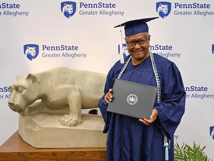 A person wearing a cap and gown in front of a Nittany Lion statue. 