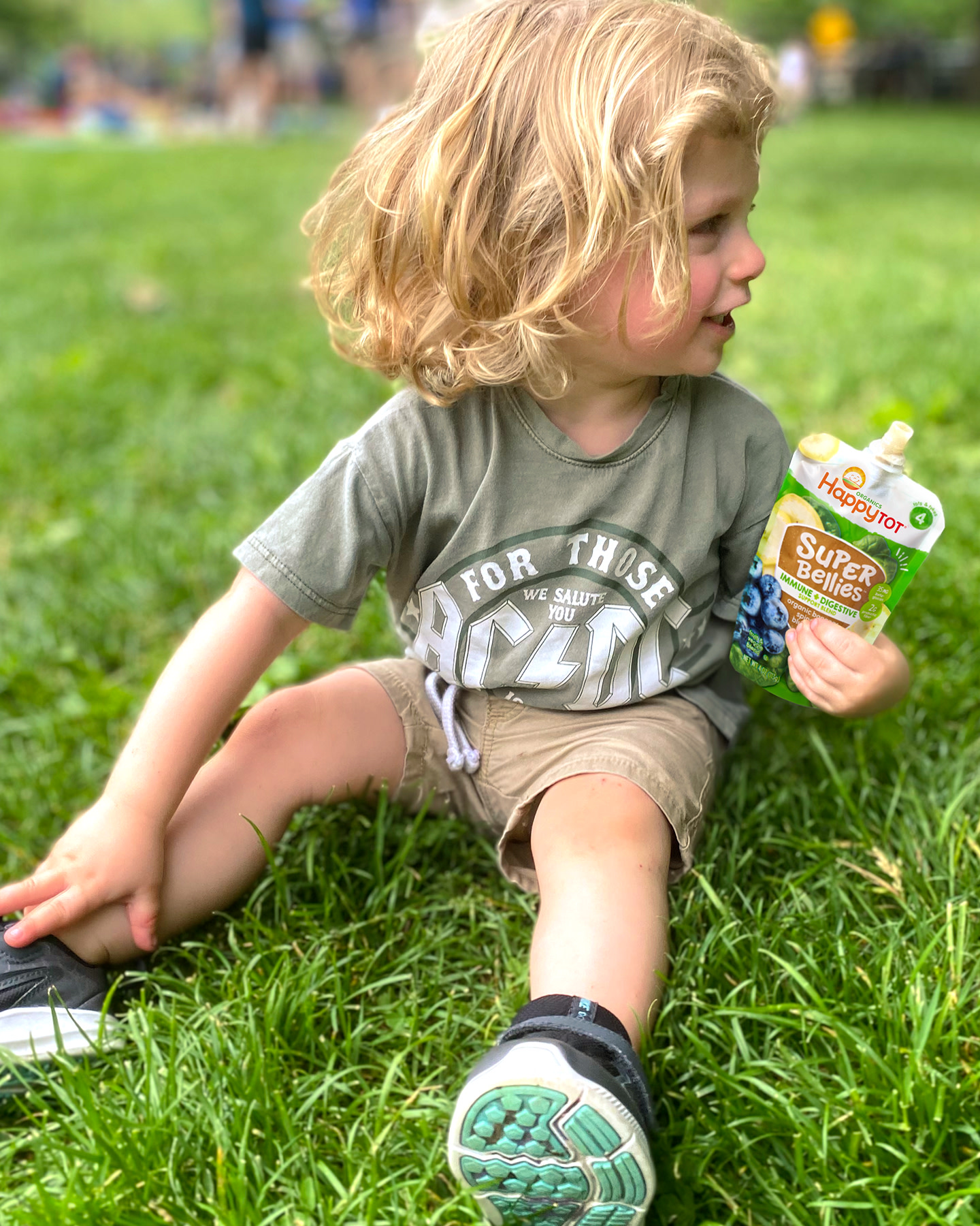 Toddler sitting on grass in park, holding up a Super Bellies pouch and smiling to the left