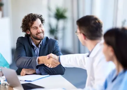 A business man in a suit shakes another man's hand at a conference table