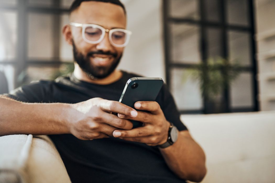 Photography of a black man with a bead and glasses relaxing on the couch, smiling while looking down at his mobile phone
