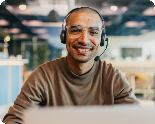 A young man in a mock turtleneck wearing a headset smiles from behind his computer screen