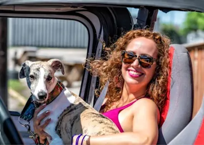 A happy woman with large curly hair and sunglasses smiles from the front seat of her Jeep with her dog in her lap and the roof off.