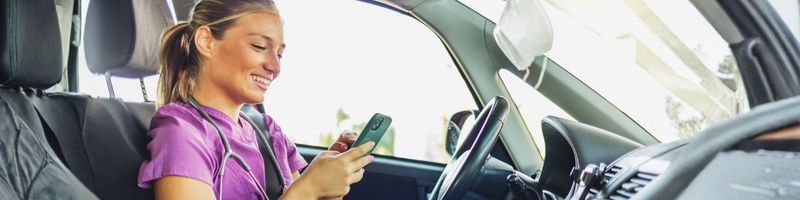 Photograph of a young female nurse wearing purple scrubs, sitting in her car using her mobile phone and smiling