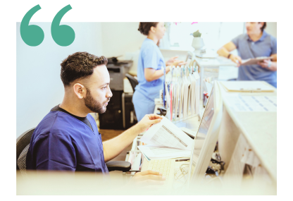 A billing coder in scrubs sits at a desk in a facility office using the computer