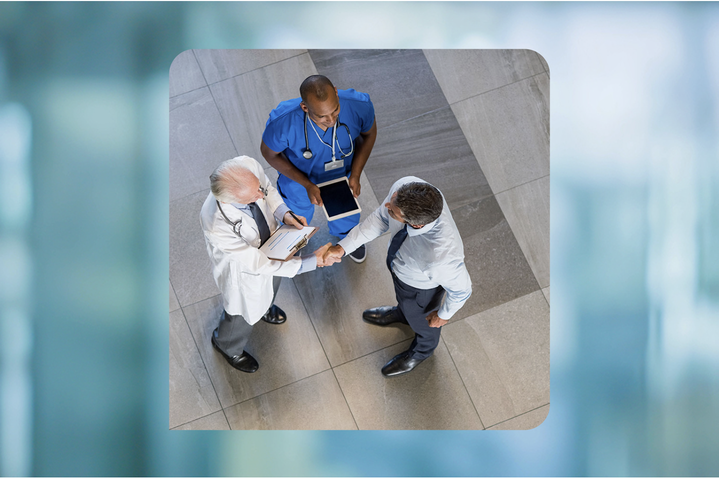 Three men meet in a facility lobby - a physician in a white coat shakes the hand of a man wearing a shirt and tie, while a man in scrubs looks on with a tablet