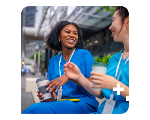 Two female healthcare professionals in scrubs sit on a bench laughing and drinking coffee