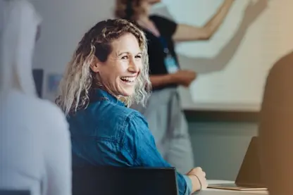 A woman in her 30s with blond hair smiles widely over her shoulder in a classroom