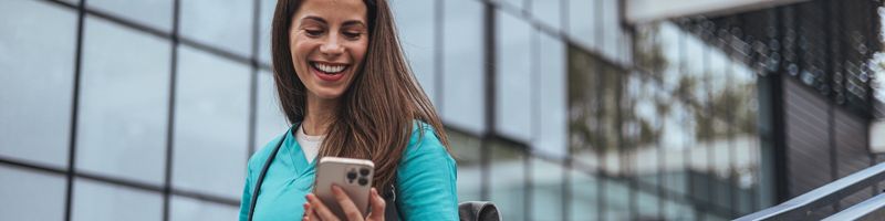 Photograph of a woman with long hair in blue scrubs walking outdoors and smiling at her mobile phone as she walks