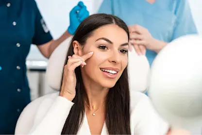 Person sits in a clinic chair holding a mirror while two medical staff stand nearby.