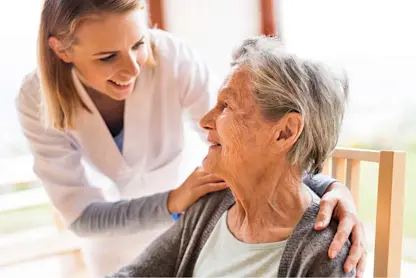 Caregiver gently supports an older adult seated in a wooden chair indoors.