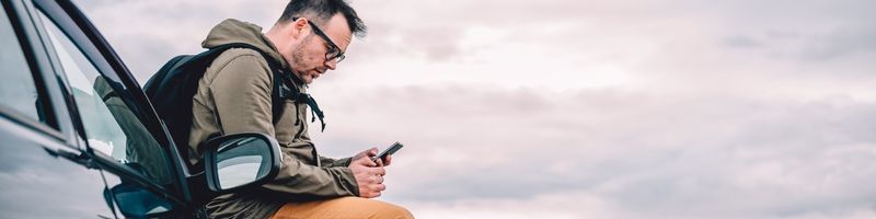 Photograph of a man outdoors, leaning on the hood of his car as he scrolls on his mobile phone